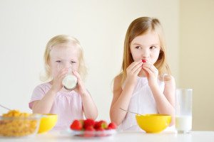 Two cute little sisters eating cereal with strawberries and drinking milk in white kitchen